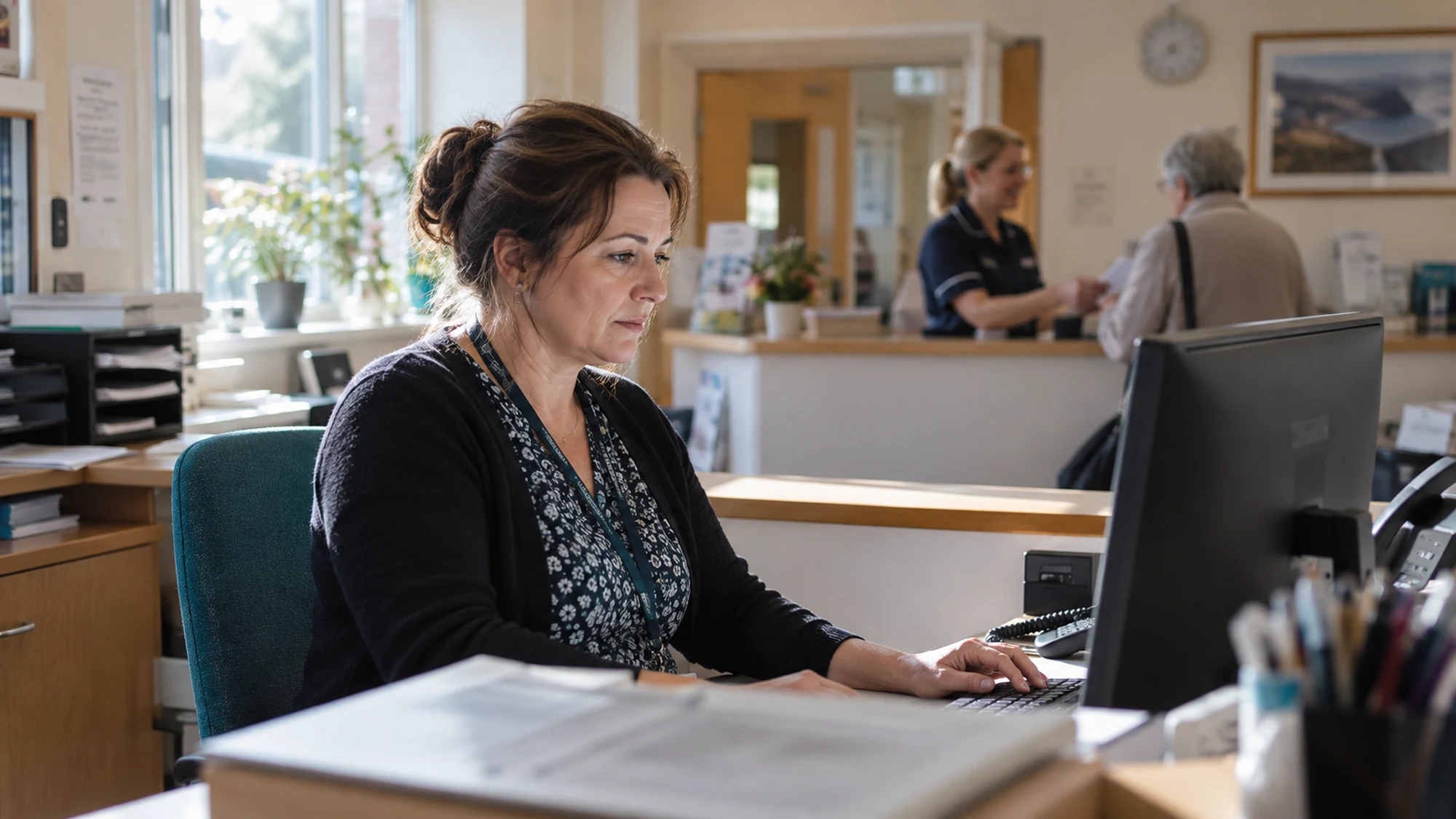 Editorial image of a practice administrator working at reception while a colleague helps a patient.
